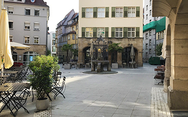 Der Hans-im-Glück-Brunnen. Im Hintergrund die Café-Bar Deli, links die Außenbestuhlung der Kneipe Platzhirsch.