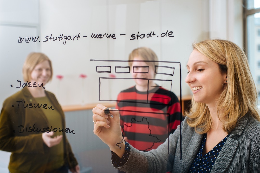 Links steht eine Frau, in der Mite ein Mann und rechts eine Frau, die auf einer durchsichtigen Plexiglasscheibe mit einem Stift eine Skizze einer Webseite anfertigt. Foto: Martin Lorenz/Stadt Stuttgart